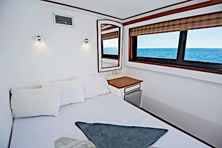 Bedroom on a boat with a white bed, white pillows and two windows showing a blue ocean