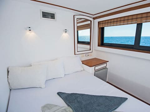 Bedroom on a boat with a white bed, white pillows and two windows showing a blue ocean