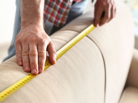 two hands holding yellow measuring tape placed on top of tan couch cushions